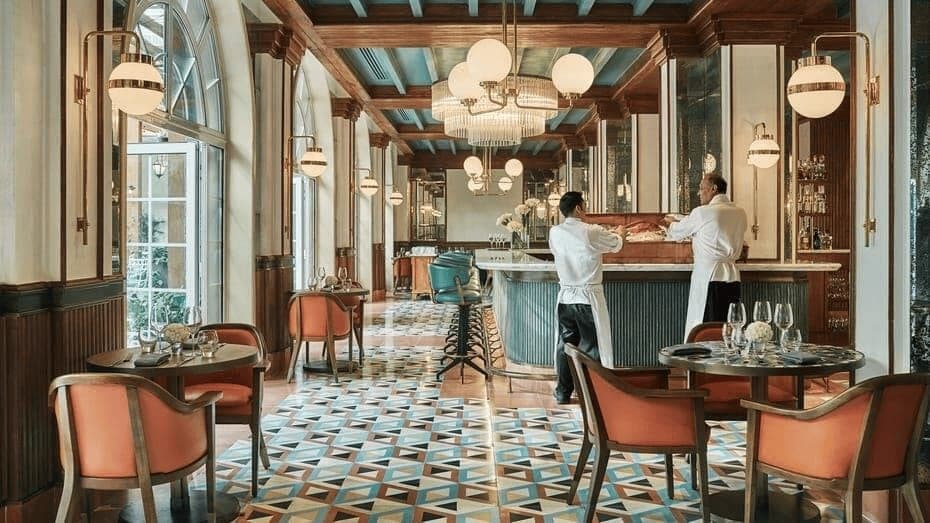 A restaurant with tiled floors, orange chairs and waiters at the bar