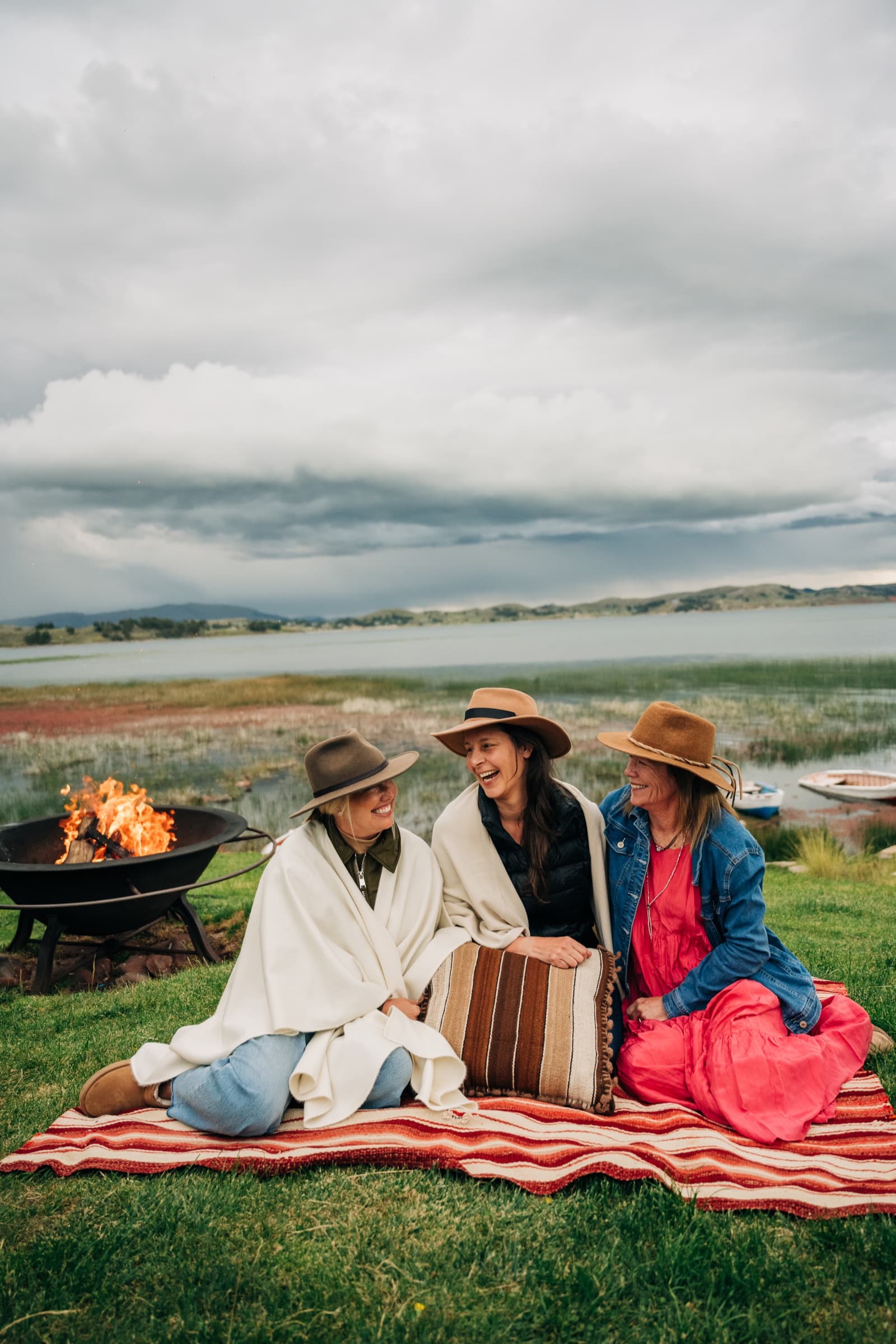 Three travelers laughing around a fire pit by a lake