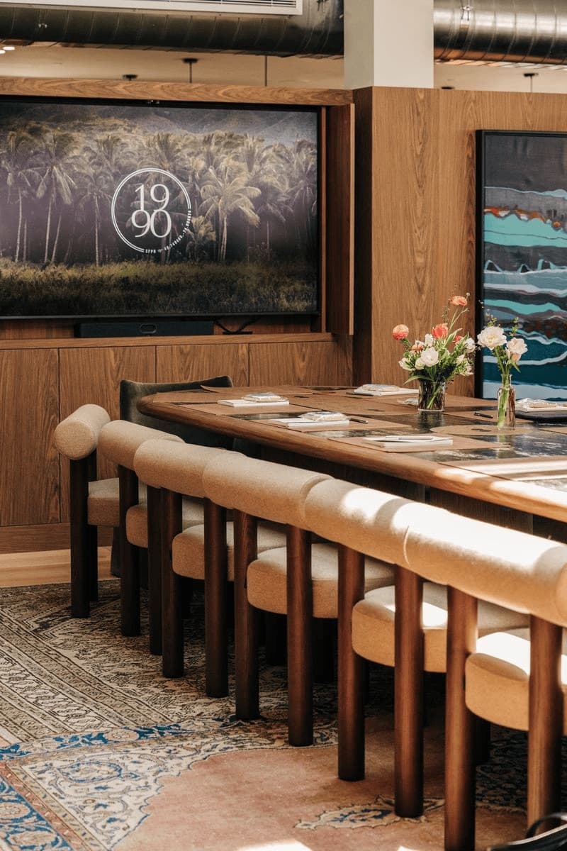 A long wooden table with flowers, upholstered chairs and a screen in a meeting room