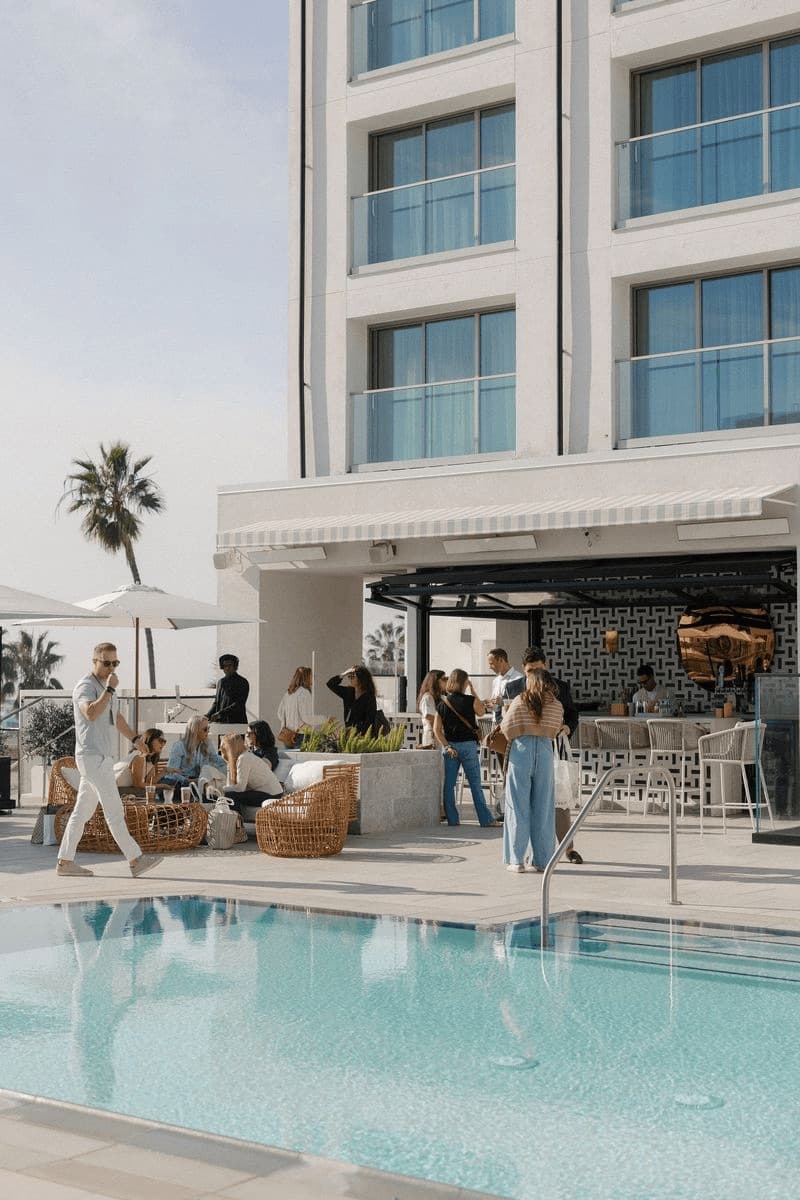 Guests around a hotel pool terrace with palm trees and a white building