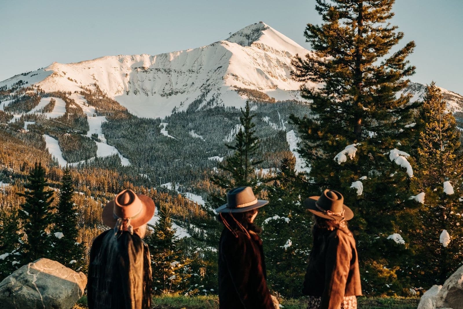 Three travellers in wide-brimmed hats looking over a snowy valley and pine forest at sunset